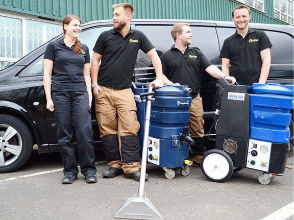 This is a photo of Mile End Carpet Cleaning carpet cleaners (three men and one woman) standing in fromt of their black van, with two steam cleaning carpet machines next to them