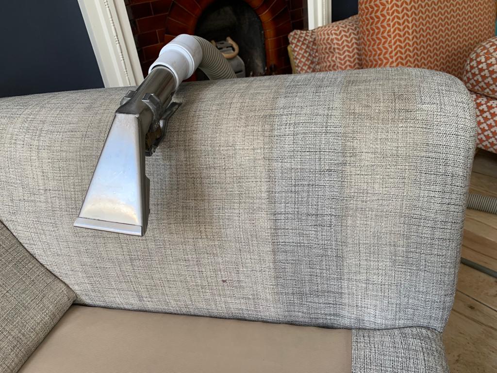 This is a photo of an arm of beige sofa that shows a test patch that has been steam cleaned. The steam cleaning machine is also showing in the photo works carried out by Mile End Carpet Cleaning